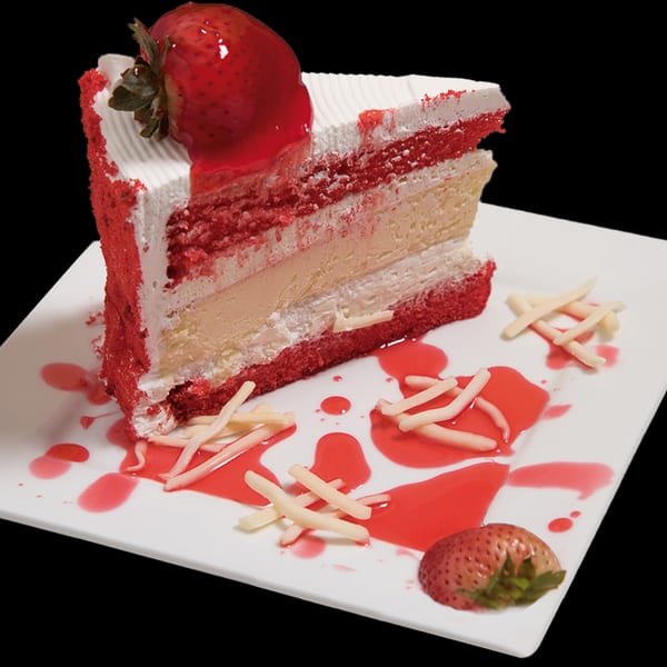 Slice of red velvet cake with white frosting, strawberry topping, and red sauce on a white plate.