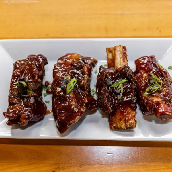 Four Jamaican jerk short ribs, glazed with sauce and garnished with herbs, on a white plate.