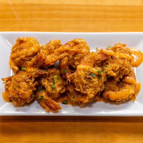Crispy fried shrimp drizzled with a creamy orange sauce and garnished with green herbs on a white plate.
