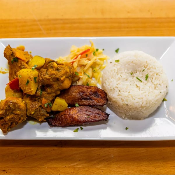 A Jamaican curry chicken dish with rice, plantains, and coleslaw on a white rectangular plate.
